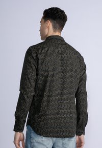 Camisa negra con un patrón geométrico dorado repetido, de mangas largas y puños abotonados. La vista trasera muestra un dobladillo curvado y un diseño de cuello.
