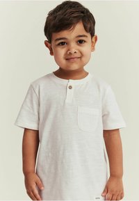 Hvid bomulds T-shirt med korte ærmer, rund hals, lomme foran og detaljer med træknapper. Blød tekstur og afslappet pasform.