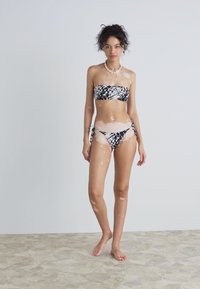 Svart og hvit mønstret bandeau-bikini med justerbare snøring i sidene. Glatt stoff med en myk glans, vist på en modell mot en lys bakgrunn.