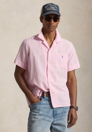 Homme portant une chemise rose à manches courtes boutonnée, un jean bleu, une casquette de baseball bleu marine, des lunettes de soleil, une ceinture marron et une montre-bracelet, debout avec une main dans la poche.