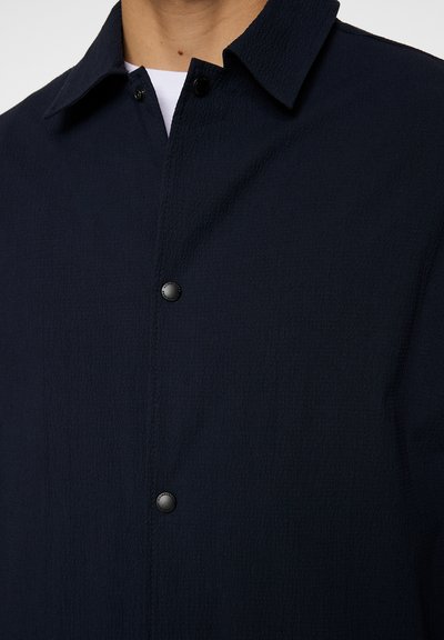 Marineblauwe button-up jas van een gestructureerde stof, met een spitse kraag en zwarte drukknoopjes, deels zichtbaar in een close-upweergave.