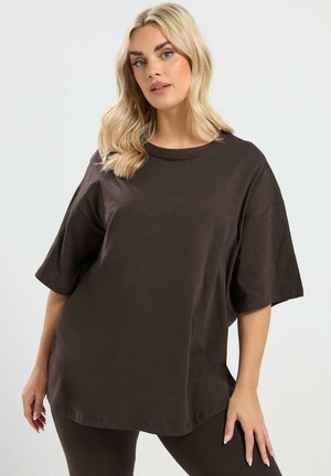 Blonde Frau, die ein übergroßes dunkelbraunes T-Shirt und passende Leggings trägt, posiert vor einem schlichten hellen Hintergrund.