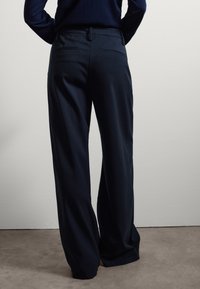 Pantalons larges bleu marine en tissu lisse, présentant une taille mi-haute, des passants de ceinture et des poches passepoilées à l'arrière. Design uni sans motifs.