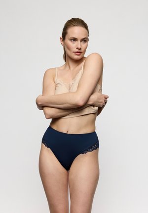 Vrouw met lichte huid en haar in een staart draagt een beige camisole en marineblauw hooggetailleerd ondergoed met kant, armen gekruist over de borst, neutrale achtergrond.
