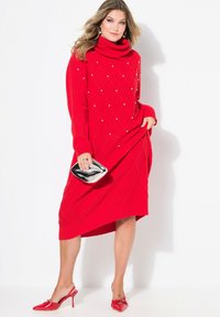 Rotes gestricktes Kleid mit hohem Kragen, das mit Rautenmuster und Perlenakzenten verziert ist, kombiniert mit einer metallischen Clutch und roten Absätzen.