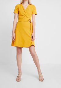 Vikklänning i klart gult tyg, med korta ärmar, en v-ringning och en midja som betonas av en cirkulär ringdetalj.