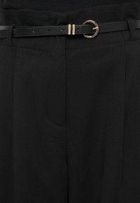Pantalon noir sur mesure avec poche avant et fine ceinture en cuir noir avec une petite boucle ronde dorée et deux passants dorés.