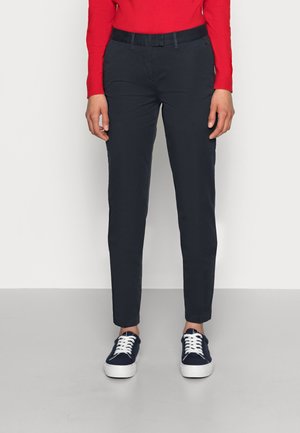 Person iført marineblå skræddersyede bukser, rød langærmet top og marineblå sneakers med hvide såler og snørebånd, stående mod en ensfarvet baggrund.