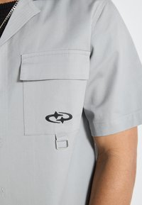 Siva majica kratkih rukava s teksturiranim završetkom, koja ima džep na prsima, crni emblem s стрелицама i detalj od petlje od hardvera.