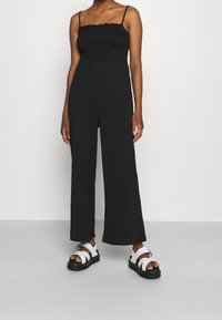 Combinaison noire avec un buste ajusté et un pantalon évasé. Présente des bretelles spaghetti et un col volanté. Associée à des sandales blanches.