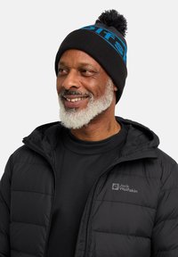 Piumino nero con cappuccio e chiusura zip, caratterizzato da un logo grigio. Berretto a maglia nero con testo blu e pompon in cima.