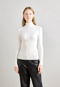 Pull en maille côtelée blanche avec une silhouette ajustée et des détails de panneaux verticaux, associé à un pantalon en cuir noir pour le contraste.