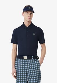 Marinblå polo skjorta med klassisk krage och logotyp, tillsammans med rutig shorts i blått och grönt. Uppsättningen har en matchande marinblå visir.