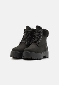 Botas cortas negras de ante, con un acabado de ante, cuello de pelaje sintético texturizado y suela de goma gruesa con patrones de tracción y cordones negros.
