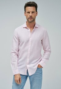 Camisa de botones de manga larga en lino color rosa claro, con cuello estándar y un único logotipo en el pecho. Combinada con jeans azules.