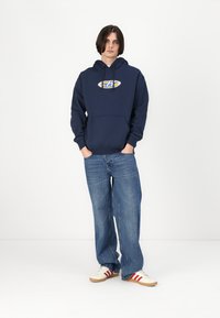 Un hoodie bleu marine avec un logo à l'avant, une capuche à cordon et une poche kangourou, associé à un jean large bleu clair et des baskets blanches.