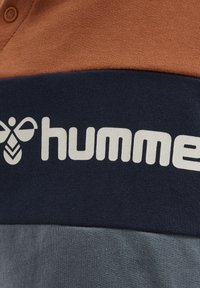 Kortärmad skjorta med en flerfärgad design i brunt, marinblått och grått. Inkluderar en framträdande vit logotyp på den marinblå sektionen.