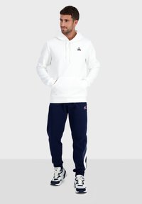 Hoodie blanc avec une poche frontale et un logo sur la poitrine, associé à un pantalon de jogging bleu marine avec des rayures latérales et des accents blancs. Baskets sportives.