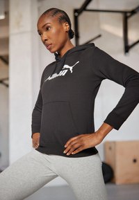 Must musta kapuutsiga särk, millel on esitasku ja valge "PUMA" logo. Kombineeritud heledate hallide retuusidega, mis on sileda tekstuuriga.