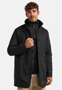 Chaqueta negra acolchada con cuello alto, cierres de botón y bolsillos laterales. Textura de tela suave y diseño elegante y moderno.