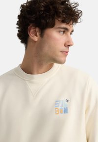 Sudadera color crema con cuello redondo y puños acanalados. Incluye un gráfico colorido con texto azul y naranja. Textura de tela suave.