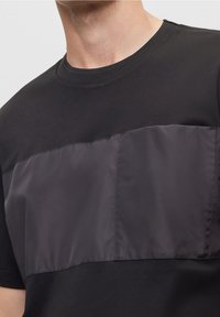 Muž v černém tričku s lesklou, hladkou vodorovnou panely přes hruď, střiženým na krku a ramenou.