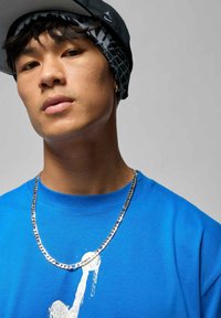 Blaues Baumwoll-T-Shirt mit weißem Aufdruck, silberne Kettenhalskette und gemustertes schwarzes und graues Bandana unter einer schwarzen Kappe. Neutraler Hintergrund.