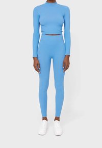Jasnoniebieski prążkowany crop top z długim rękawem w zestawie z dopasowanymi legginsami o wysokim stanie. Noszony z białymi trampkami, minimalistyczny design, gładka tekstura.