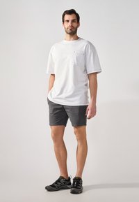 Camiseta de algodón blanca con un bolsillo en el pecho, combinada con pantalones cortos grises. El modelo lleva zapatillas deportivas negras con detalles verdes.