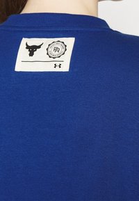 Blå bomullströja med ribbat rundhals. Har en vit etikett på ryggen som visar en tjur-symbol och varumärkesdetaljer.