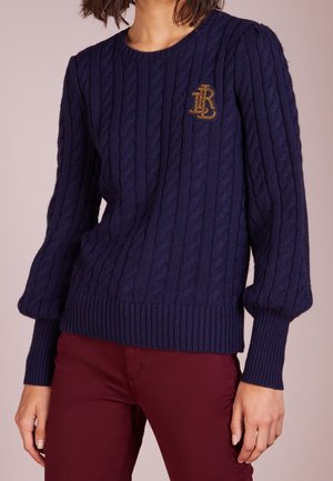 Granatowy sweter w warkoczowy splot z złotym haftowanym logo na piersi, zestawiony z bordowymi spodniami.