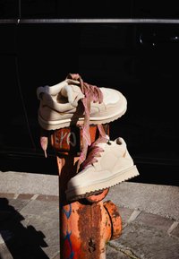 Béžové tenisky s texturovanou tkaninou, růžovými šňůrkami s vzorem a silnou gumovou podrážkou, opřené o rezavě oranžový hasičský hydrant.