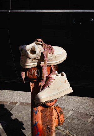 Béžové tenisky s texturovanou tkaninou, růžovými šňůrkami s vzorem a silnou gumovou podrážkou, opřené o rezavě oranžový hasičský hydrant.