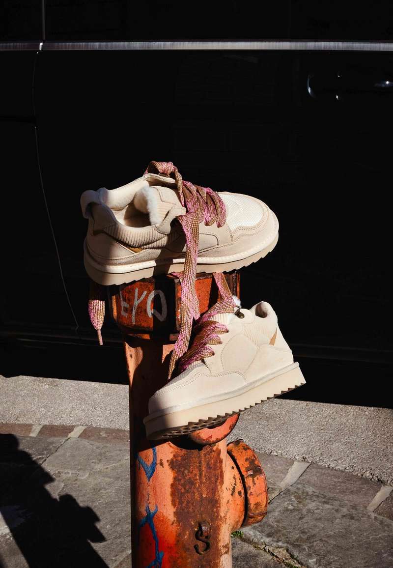 Béžové tenisky s texturovanou tkaninou, růžovými šňůrkami s vzorem a silnou gumovou podrážkou, opřené o rezavě oranžový hasičský hydrant.
