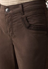 Gros plan d’un pantalon marron foncé montrant une poche avant avec bordure texturée, passant de ceinture, fermeture par bouton et une partie d’un pull en tricot beige.