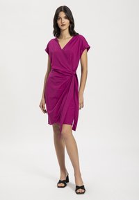 Fukso wrap-dressillä on lyhyet hihat, drapeerattu etuosa ja sivusolmu. Sujuva materiaali ja rento malli, yhdistettynä mustiin paljaisiin korkokenkiin.