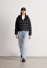 Czarna kurtka puchowa z zamkiem z przodu, regulowanym kapturem i teksturowanym wykończeniem. Połączona z jasnoniebieskimi luźnymi dżinsami i czarnym obuwiem.
