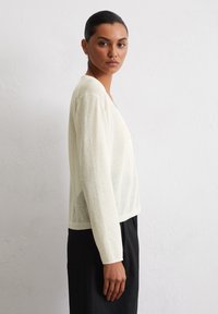 Leichte, durchsichtige elegante Cardigan mit langen Ärmeln in einem sanften Gelbton, mit V-Ausschnitt, subtiler Textur und lässiger Passform, getragen über einem schwarzen Outfit.