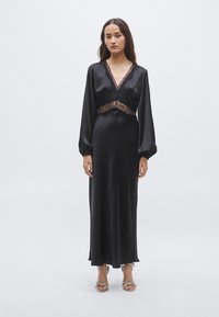 Čierne saténové šaty s čipkovými detailmi, dlhými rukávmi, hlbokým výstrihom a zvýrazneným pásom. Sukňa splýva až k členkom so subtílnym leskom.