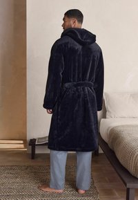 Navy blue plush bathrobe with a hood, tied at the waist, worn over light gray striped pants; soft texture against a neutral background.