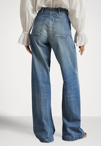 Γυναίκα που φορά φαρδιά μπλε τζιν παντελόνια και λευκό μπλουζάκι με μακριά μανίκια και σούρες στα μανίκια, εμφανιζόμενη από πίσω.