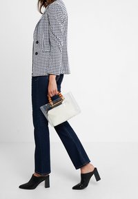 Blazer houndstooth bianco e nero, jeans scuri e mule nere con tacco alto; tiene in mano una borsa trasparente con un sacchetto bianco e manico in legno.