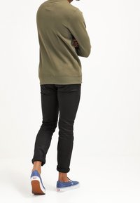 Sudadera verde oliva, vaqueros ajustados negros y zapatos de lona azules con detalles blancos y suelas de goma. De pie, vista desde atrás.