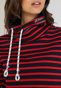 Felpa a strisce rossa e blu navy con colletto alto, dotata di lacci bianchi con dettagli neri e una zip argentata sul collo.