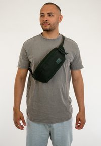 Borsa a tracolla verde con forma rettangolare, realizzata in tessuto. Presenta una chiusura con zip e una tracolla regolabile per indossarla a tracolla.