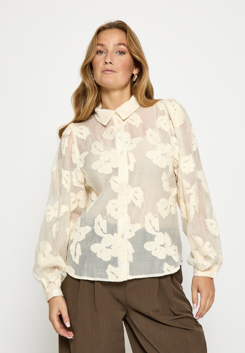 Przezroczysta beżowa bluzka w kwiatowy wzór, z guzikami z przodu, kołnierzykiem i długimi bufkowatymi rękawami. Tkanina o strukturze z delikatnym połyskiem.