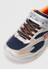 De Skechers S-Lights sneaker heeft een marineblauwe mesh stof, zilveren synthetische accenten, oranje veters en een gestructureerde zool met verlichting.