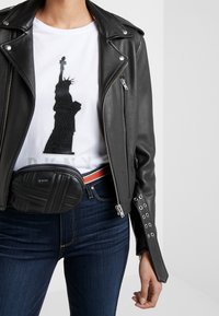 Personne portant une veste en cuir noire, un T-shirt blanc avec un motif de la Statue de la Liberté, un jean sombre et une sacoche ceinture en cuir noir avec une ceinture à rayures rouges.