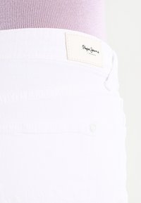 Bílé džínové šortky s texturovaným povrchem, s výraznou značkovou štítkem na pásku a jedním kovovým tlačítkem.