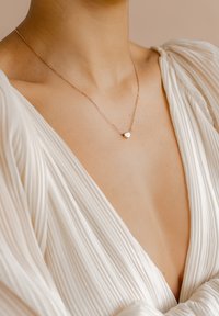 Collana dorata con un piccolo pendente a forma di cuore, posata su un collo nudo, caratterizzata da un sottile design a catena curb e una texture delicata.